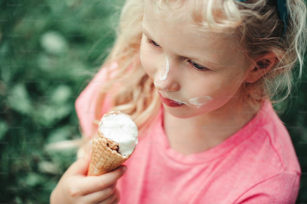Cute funny adorable girl with dirty nose and milk moustaches eating licking ice cream from waffle cone. Child eating tasty sweet cold summer food outdoors. Summer frozen meal snack.