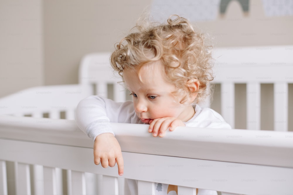 Lonely thoughtful baby boy toddler standing in crib at kids nursery room at home. Sad pensive thinking baby boy with curly blond hair looking down. Authentic candid home life childhood.