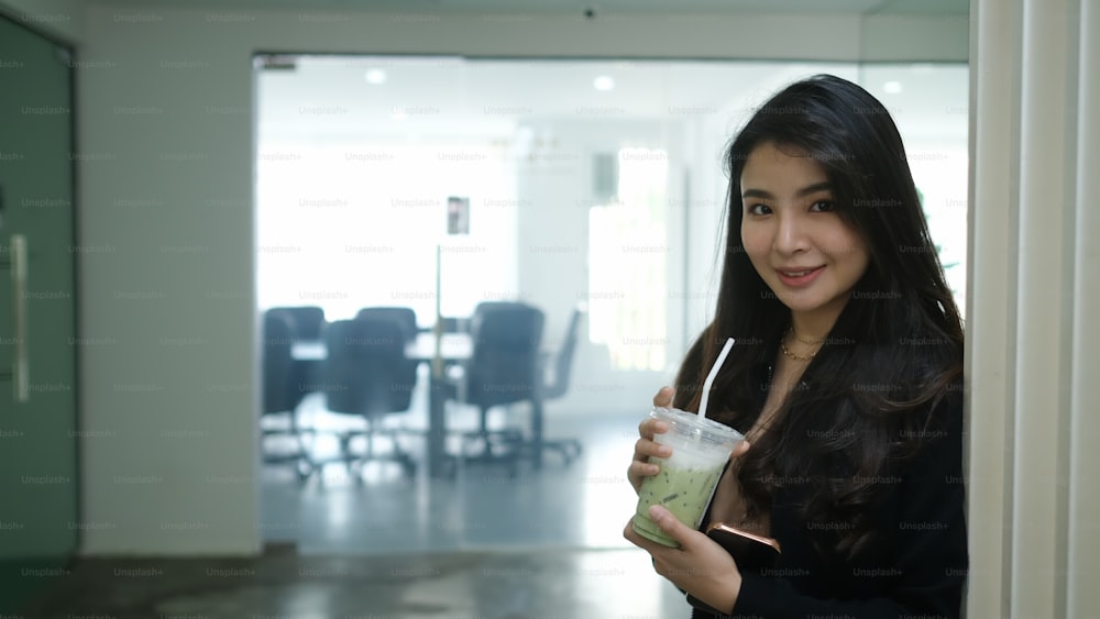Attractive businesswoman holding plastic glass of iced coffee and smiling to camera while standing in office.