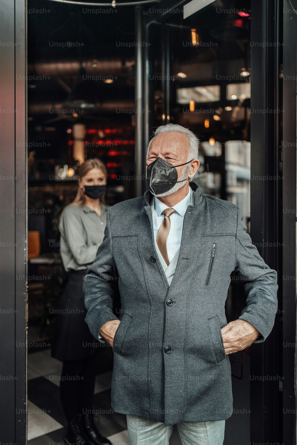 Ein älterer Geschäftsmann verlässt ein Restaurant. Junge Kellnerin verabschiedet ihn freundlich. Sie tragen eine Schutzmaske als Schutz vor einer Viruspandemie.