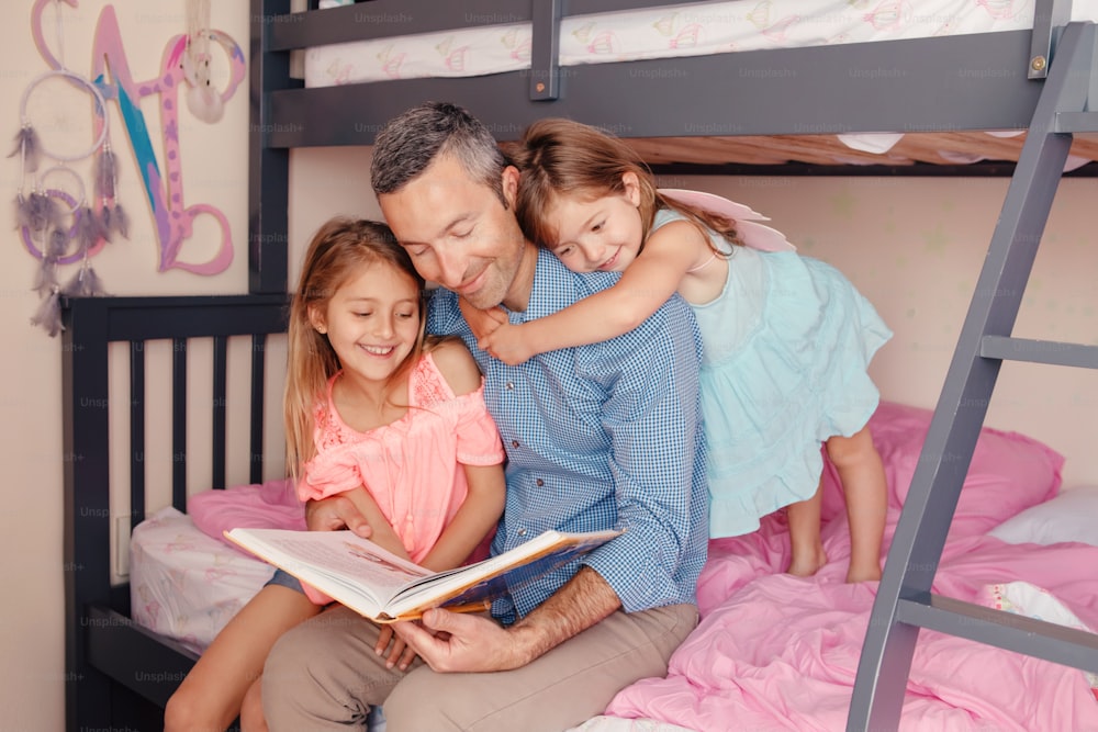 Papá leyendo un libro a las hijas. Familia de tres personas sentadas en la cama en el dormitorio. Padre e hijos felices en casa pasando tiempo juntos. Educación y un estilo de vida feliz en la infancia.