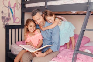 Dad reading book to daughters girls. Family of three people sitting on bed in bedroom. Happy father and children at home spending time together. Education and happy lifestyle childhood.