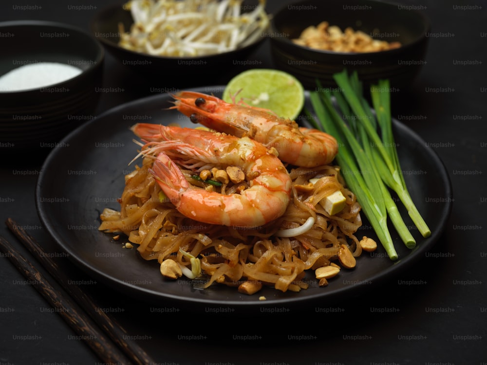 Chupito recortado de fideos tailandeses cocidos con camarones servidos en un plato negro con lima, brotes de soja y cebollino, Pad Thai