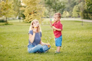 Young Caucasian mother and boy toddler son blowing soap bubbles in park. Mom and child playing having fun together outdoor on summer day. Happy authentic family childhood lifestyle.