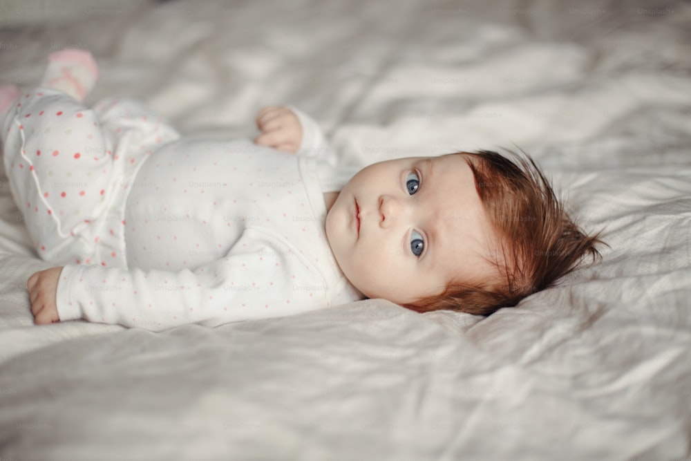 Closeup portrait of cute Caucasian newborn baby. Adorable funny child infant with blue grey eyes and red hair lying on bed looking at camera. Authentic childhood and lifestyle candid moment.