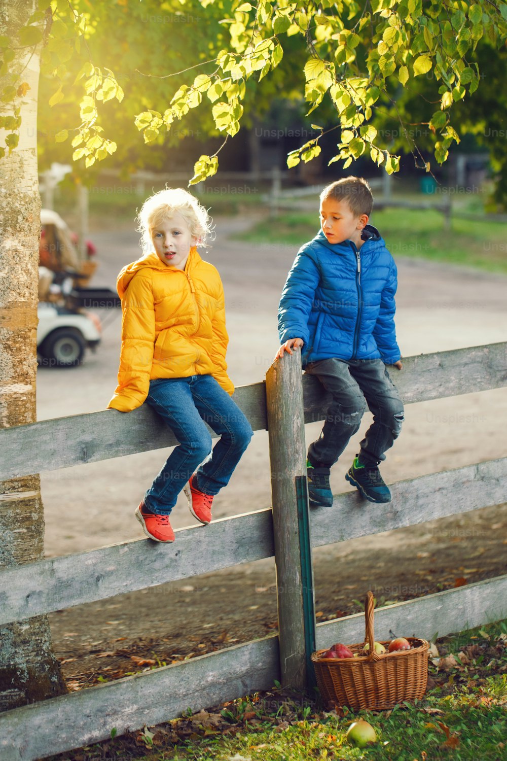Two cute funny Caucasian children boy girl sitting on wooden rustic fence in park farm outside. Friends siblings kids talking at summer sunset in countryside. Romantic love and friendship.