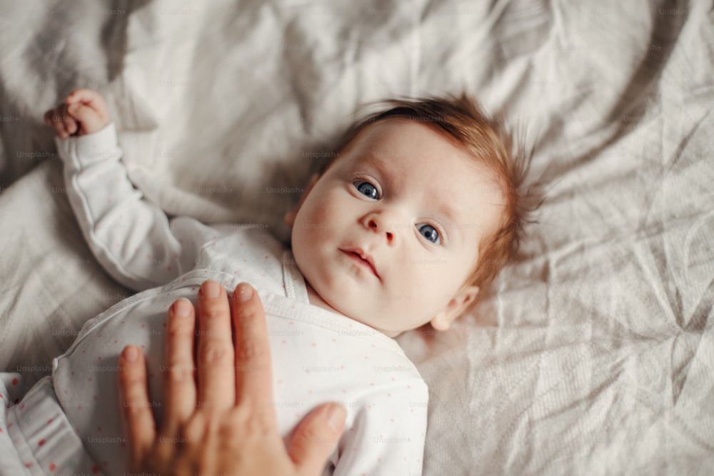 Retrato em close-up do bebê recém-nascido caucasiano bonito. Adorável criança engraçada bebê com olhos cinzentos azuis e cabelos vermelhos deitados na cama olhando para a câmera. Autêntica infância e estilo de vida momento sincero.