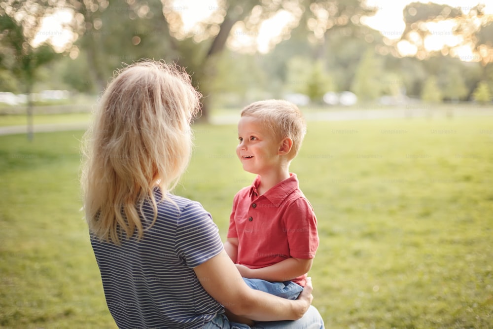 Young Caucasian mother and boy toddler son sitting together face to face. Family mom and child talking communicating outdoor on summer day. Happy authentic family childhood lifestyle.