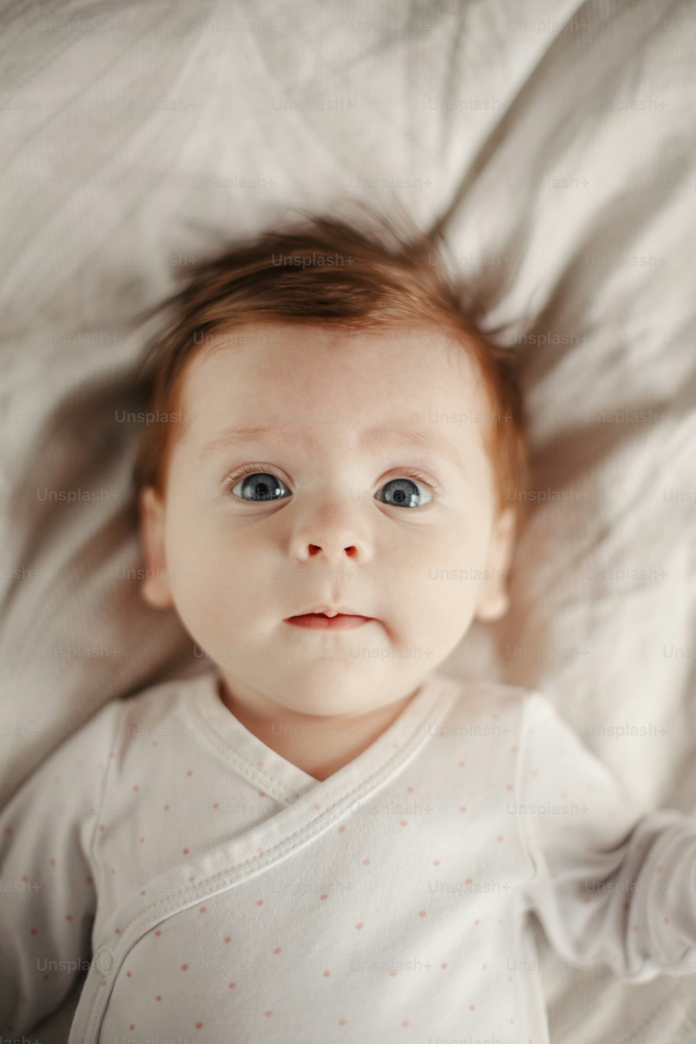 Closeup portrait of cute Caucasian newborn baby. Adorable funny child infant with blue grey eyes and red hair lying on bed looking at camera. Authentic childhood and lifestyle candid moment.