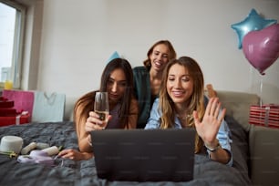Woman with a champagne flute and her two smiling friends staring at the laptop screen