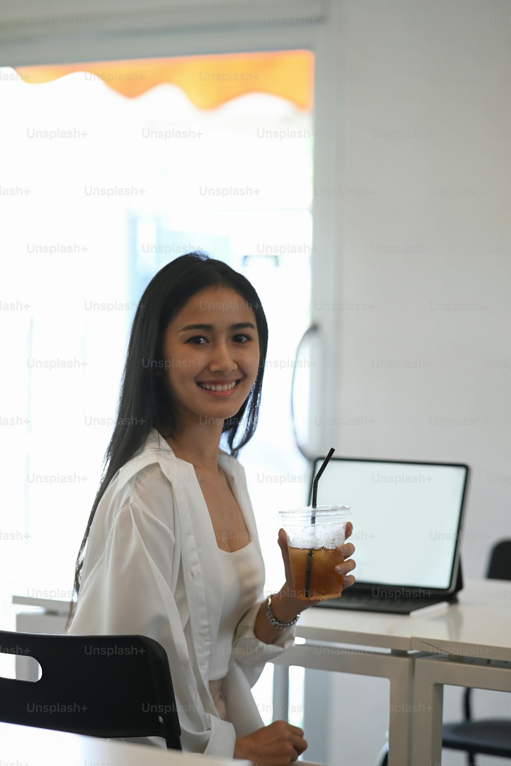 Portrait of young woman holding ice coffee cup and smiling to camera while sitting at her workspace.