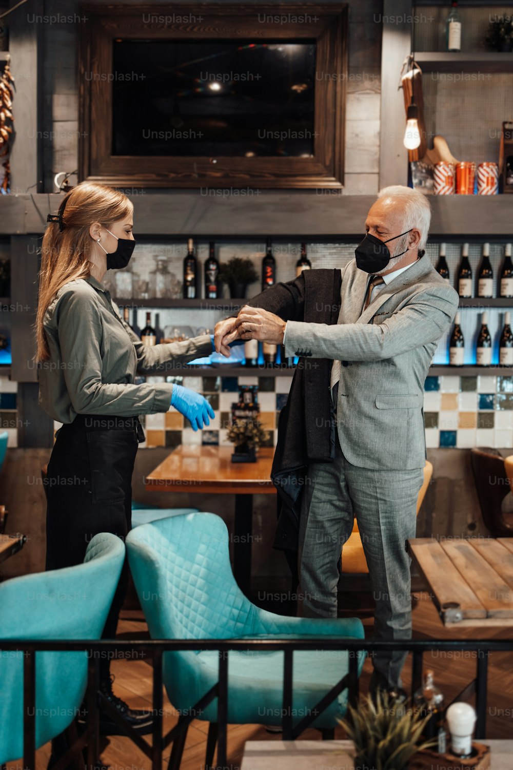 Confident senior businessman standing in exclusive restaurant. He takes off his coat while the waitress helps him. They both wearing protective face masks against virus infection.