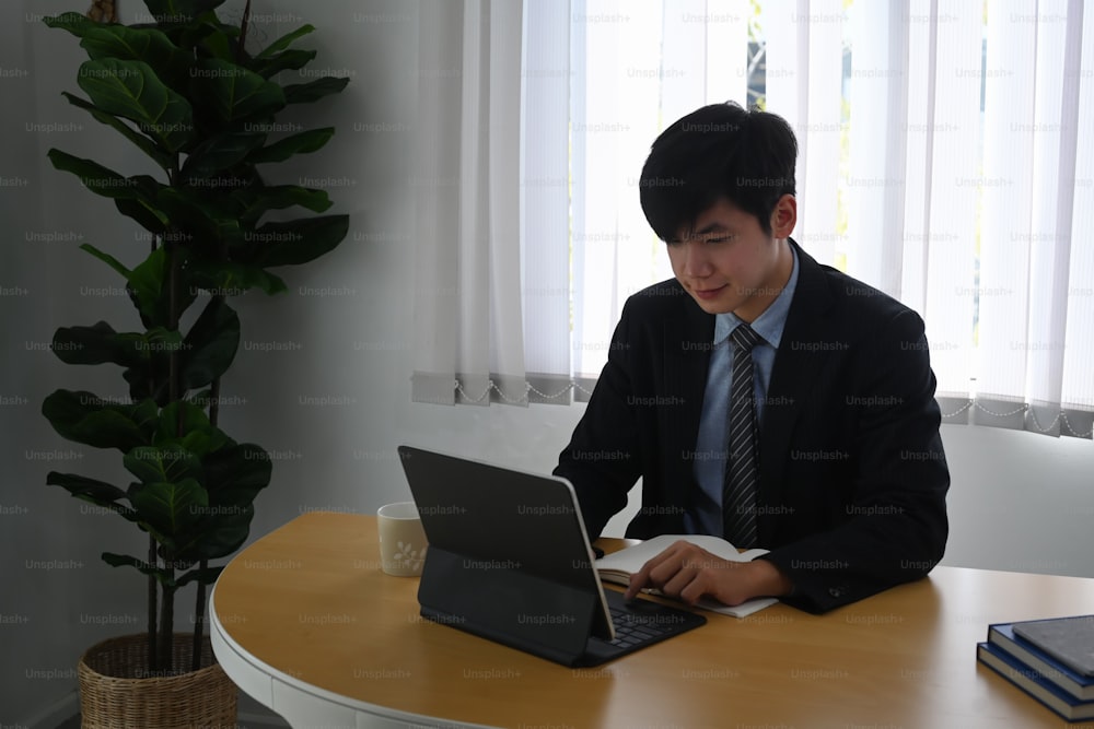 Empresário atraente trabalhando em tablet de computador e pensando em seus planos conectados com novos projetos em escritório moderno.