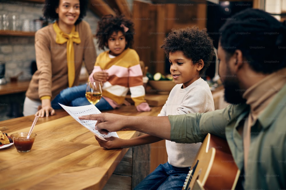 African American father explaining sheet music to his son while playing acoustic guitar at home. Focus is on boy. Mother and daughter are in the background.