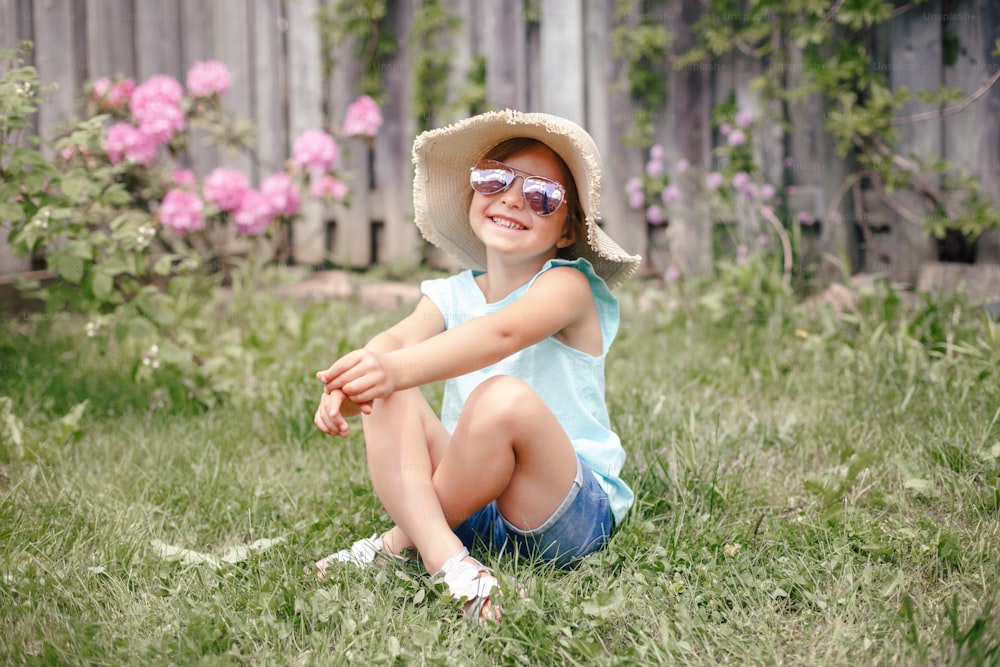 屋外の芝生の上に座っているサングラスと麦わら帽子をかぶったかわいい愛らしい子供の女の子の肖像画。自宅の裏庭で楽しんでいる幸せな笑顔の白人の子供。驚くほど楽しい夏とライフスタイルの子供時代。