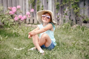 Portrait of cute adorable child girl in sunglasses and straw hat sitting on grass outdoor. Happy smiling Caucasian kid having fun at home backyard. Amazing joyful summer and lifestyle childhood.