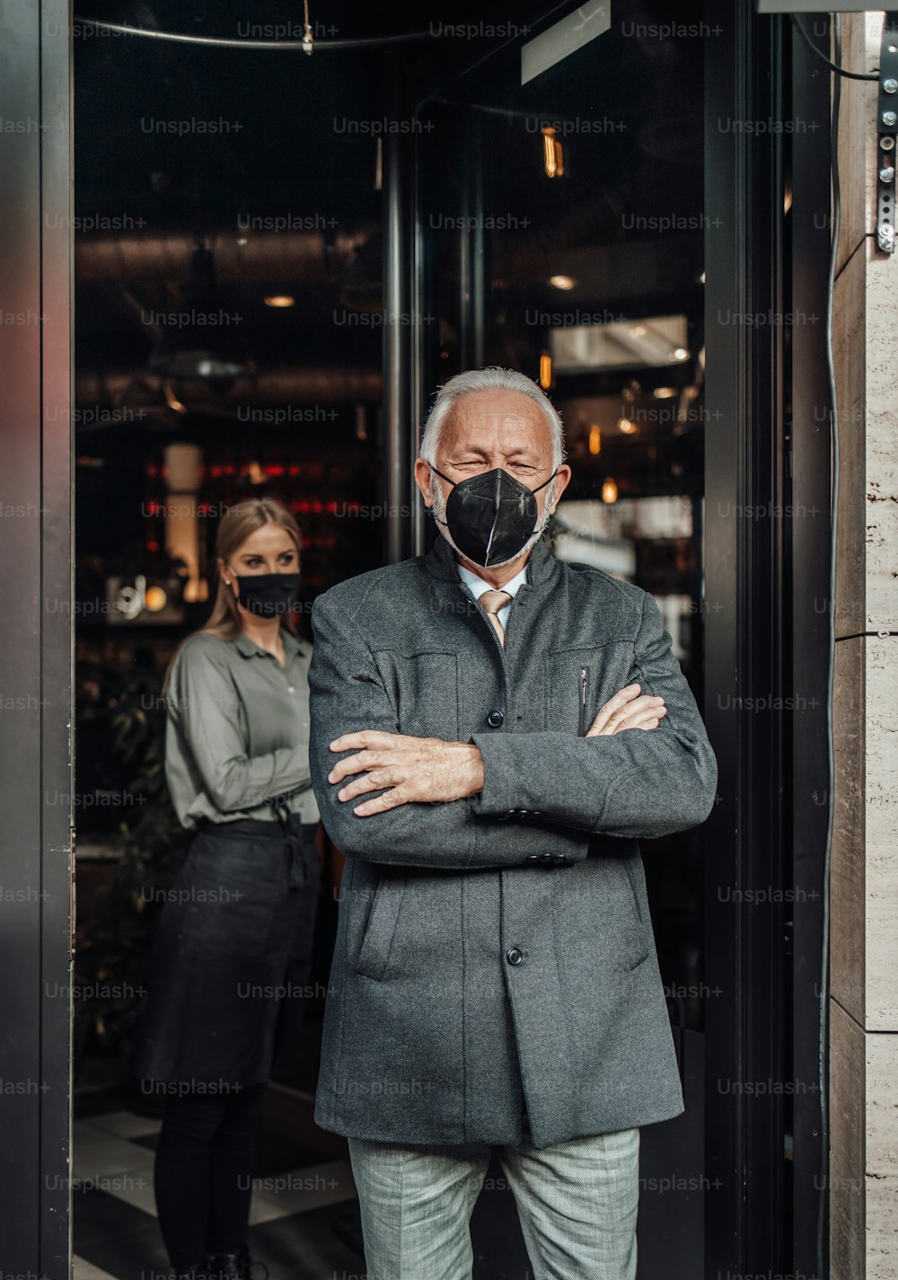 Ein älterer Geschäftsmann verlässt ein Restaurant. Junge Kellnerin verabschiedet ihn freundlich. Sie tragen eine Schutzmaske als Schutz vor einer Viruspandemie.