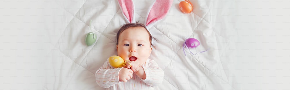 Cute adorable Asian baby wearing pink Easter bunny ears. Infant kid lying on bed with colored Easter eggs. Funny child celebrating traditional Christian holiday. Banner header for website.