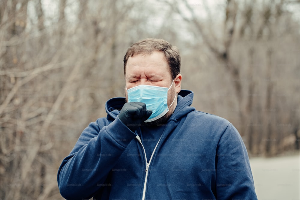 Giovane uomo di mezza età caucasico in maschera sanitaria starnutisce tosse all'aperto. Persona che protegge dalla pericolosa diffusione del virus. Quarantena della malattia da Coronavirus COVID-19.
