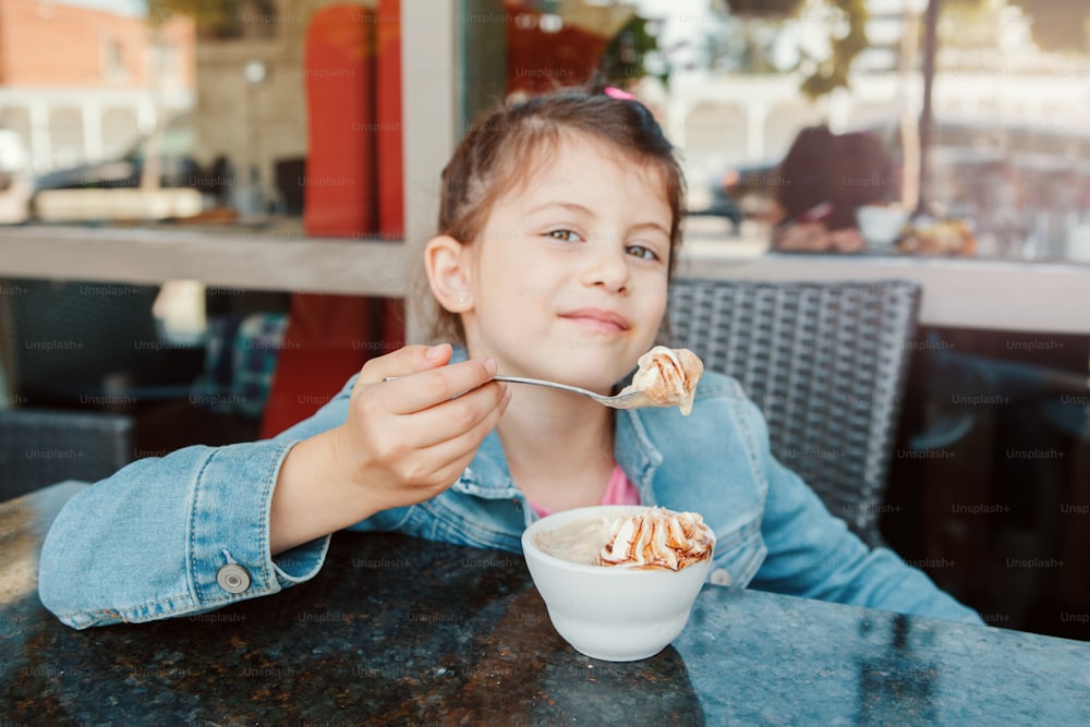 Funny Caucasian preschool girl eating sweet dessert with spoon in cafe. Child kid having fun in restaurant patio enjoying food drink. Happy authentic childhood lifestyle.