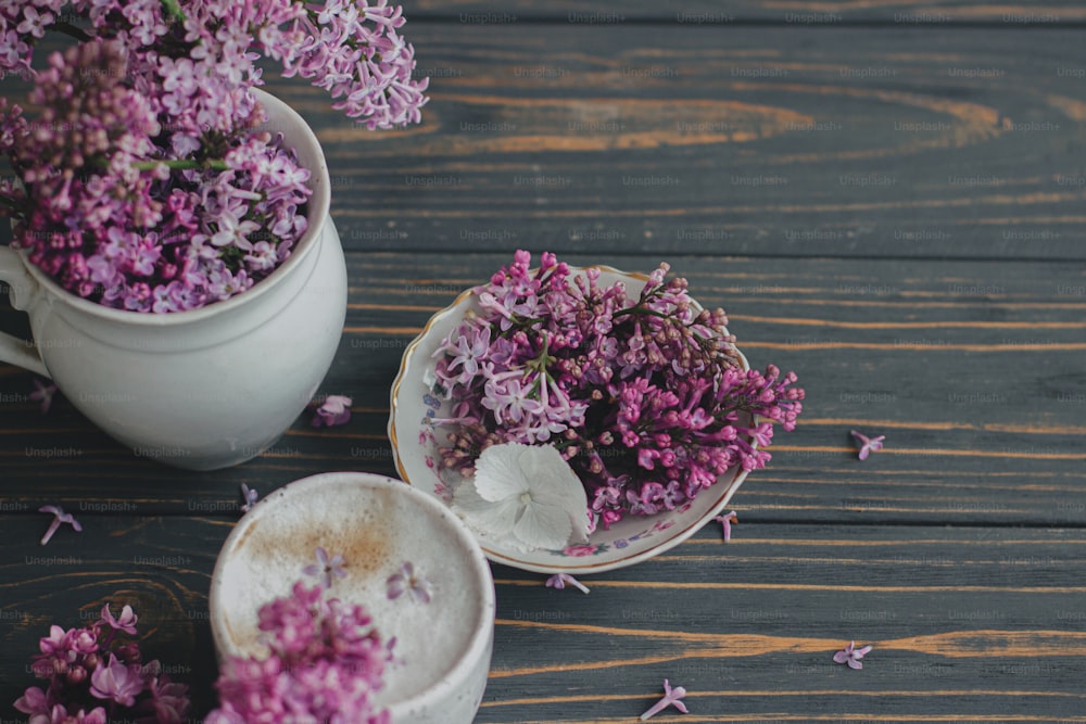 Coffee with lilac petals, blooming lilac branches in vintage cup on dark wooden background. Fresh aromatic coffee in stylish cup and spring flowers on rustic wood. Morning rituals