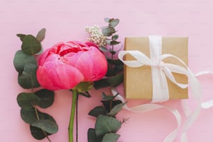 Modern minimal bouquet and simple gift box with ribbon on bright pink background flat lay. Stylish colorful greeting card with peony and eucalyptus. Happy womens day or mothers day.