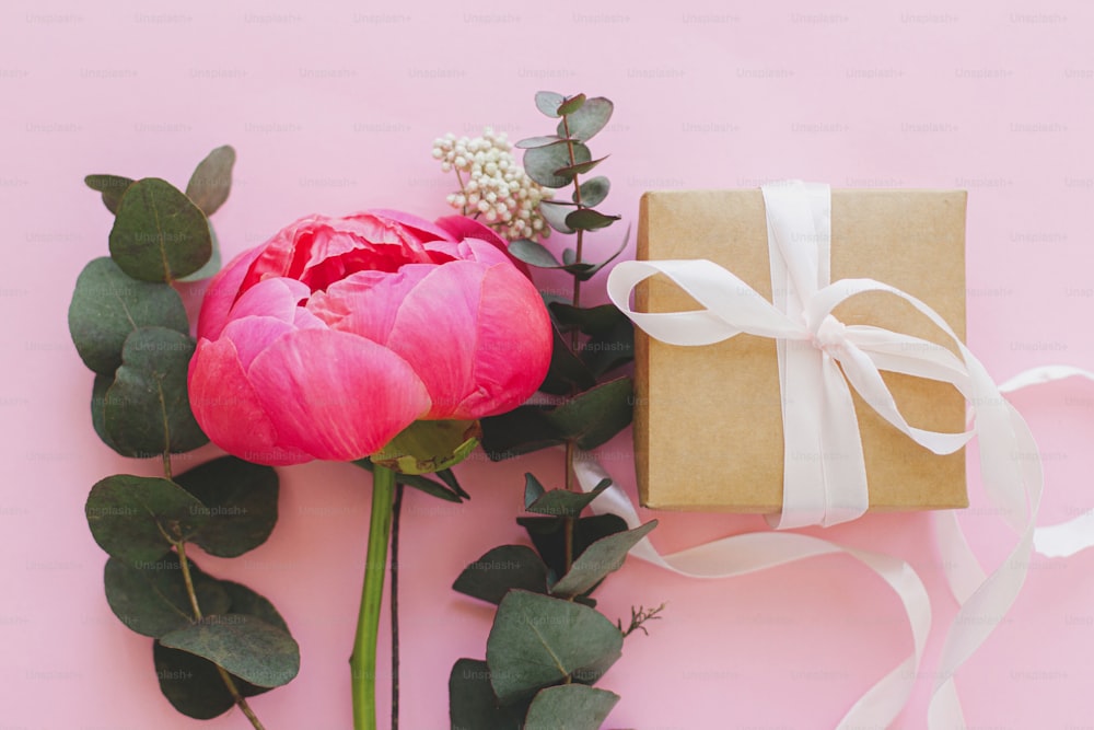 Modern minimal bouquet and simple gift box with ribbon on bright pink background flat lay. Stylish colorful greeting card with peony and eucalyptus. Happy womens day or mothers day.