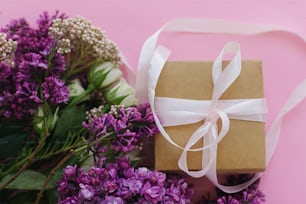 Simple gift box with ribbon and beautiful lilac and roses bouquet flat lay on bright pink background. Happy mothers day or womens day, stylish colorful greeting card.