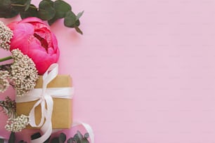 Modern fresh bouquet and simple gift box with ribbon on bright pink background,  flat lay with copy space. Happy mothers day or womens day, stylish colorful greeting card. Peony and eucalyptus