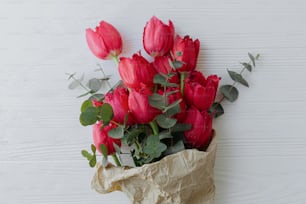 Stylish red tulips and eucalyptus bouquet in craft paper on rustic white wooden background, top view. Happy Mothers day. Happy Women's day.