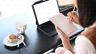 Cropped shot of young woman sitting sitting in front of her computer tablet and making notes to her diary.