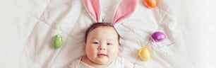 ピンクのイースターバニーの耳を身に着けているかわいい愛らしいアジアの赤ちゃん。色付きのイースターエッグでベッドに横たわっている幼児。伝統的なキリスト教の祝日を祝うおかしな子供。Web サイトのバナー ヘッダー。
