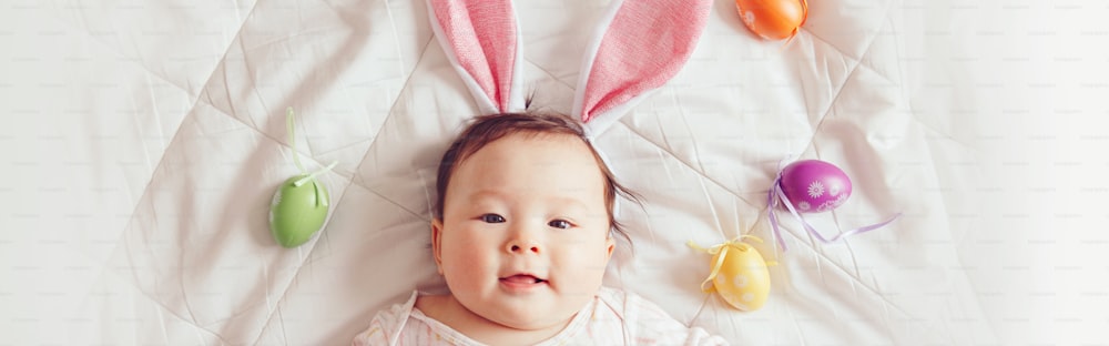 Lindo y adorable bebé asiático con orejas de conejo de Pascua rosas. Niño pequeño acostado en la cama con huevos de Pascua de colores. Niño divertido que celebra la fiesta cristiana tradicional. Encabezado de banner para sitio web.