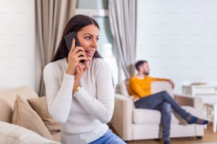 Woman turned her back to man, talking on phone with her lover, boyfriend sitting in the back watching tv. Cheating and infidelity concept