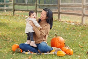 Felice madre cinese asiatica sorridente con un bambino adorabile carino. Famiglia della ragazza della mamma e della figlia che si siede nel parco autunnale all'aperto con le zucche. Concetto stagionale di Halloween o del Ringraziamento.
