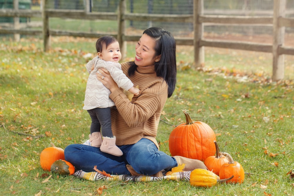 Happy smiling Asian Chinese mother with cute adorable baby. Mom and daughter girl family sitting in autumn fall park outdoor with pumpkins. Halloween or Thanksgiving seasonal concept.