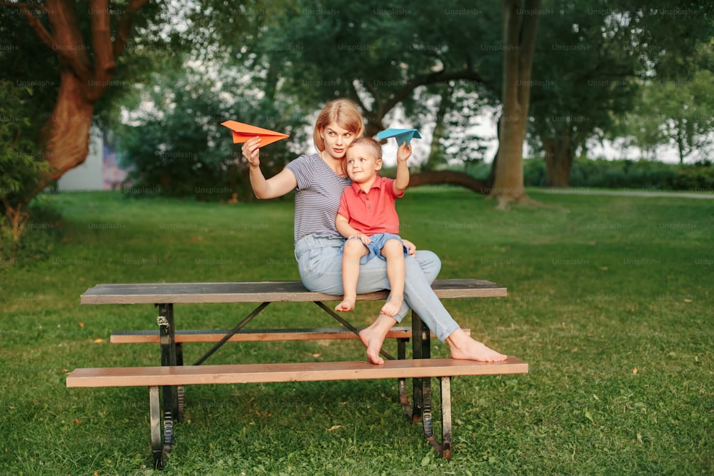 Mama und Kind spielen mit Papierflugzeugen. Junge kaukasische Mutter und kleiner Sohn sitzen am Sommertag im Freien zusammen. Glückliche Familie Kindheit Lebensstil. Menschen, die von Zukunft träumen.