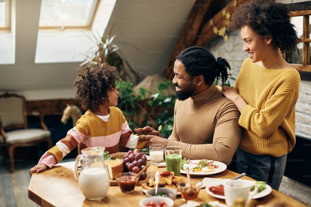 Padres negros felices y su hija pequeña comunicándose durante una comida en el comedor.