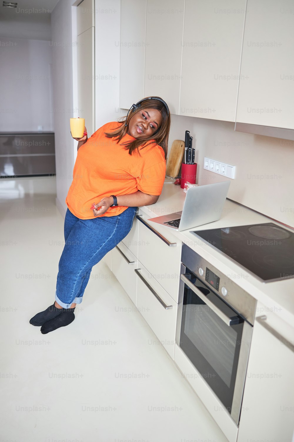 Relaxed plus-size lady smiling and drinking tea while leaning on kitchen counter and looking at laptop screen