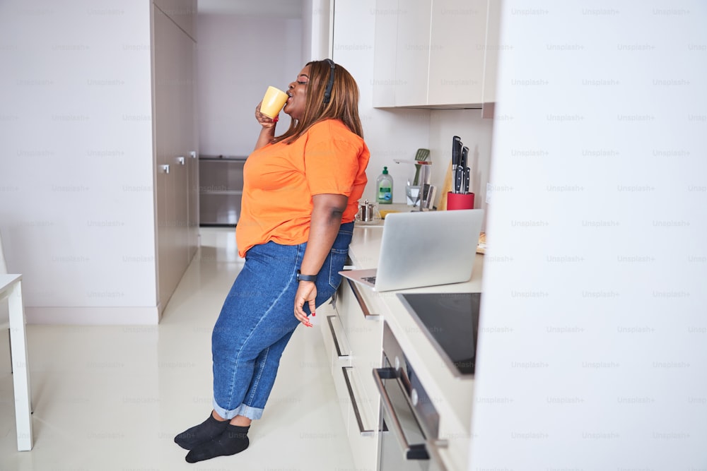 Foto de cuerpo entero de una mujer afroamericana segura de sí misma bebiendo té y de pie en la cocina