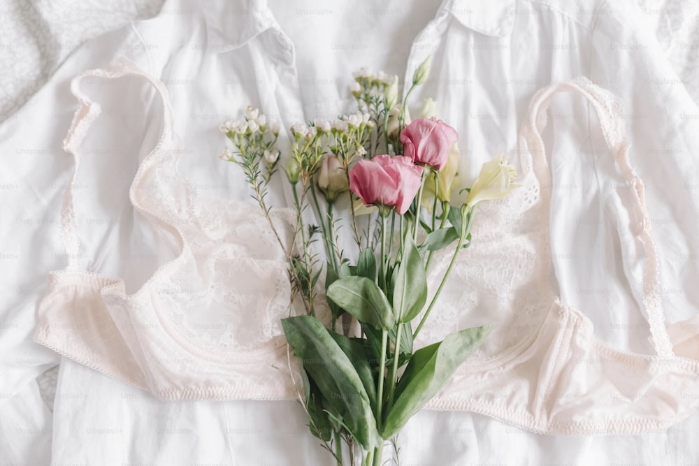 Stilvolle Spitzendessous und Frühlingsblumen auf Hemd auf dem Bett. Weiches trendiges feminines Bild, sinnliche Stimmung zu Hause Konzept. Frohen Frauentag. Damen-Essentials, Duft