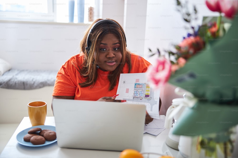 Expressive young Afro-American lady looking at laptop screen while wearing a headset and sitting in her kitchen