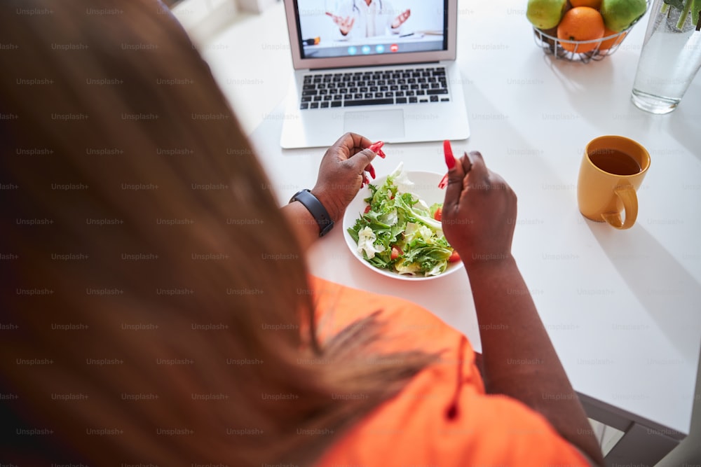 Cropped photo of ample-bodied Afro-American woman enjoying her salad while watching video on laptop
