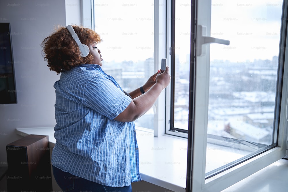 Waist-up photo of a ample-bodied Afro-American woman in headphones taking photograph of view out of her window