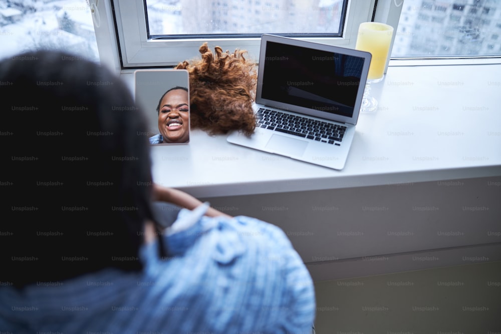 창문 근처에 앉아 거울에 비친 자신의 모습을 바라보는 즐거운 아프리카계 미국인 여성의 복사 공간 사진