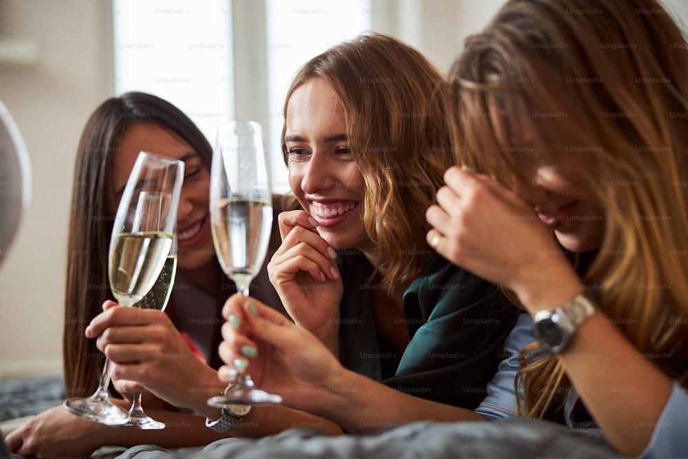 Loira feliz de boa aparência Caucasian aniversariante piscando copos de vinho espumante com duas fêmeas jovens