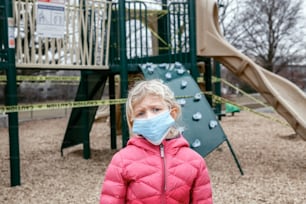 Retrato da triste menina caucasiana em máscara facial no playground fechado ao ar livre. Área de recreação infantil trancada com fita amarela de advertência em Toronto, Canadá. Quarentena de distanciamento social do coronavírus.