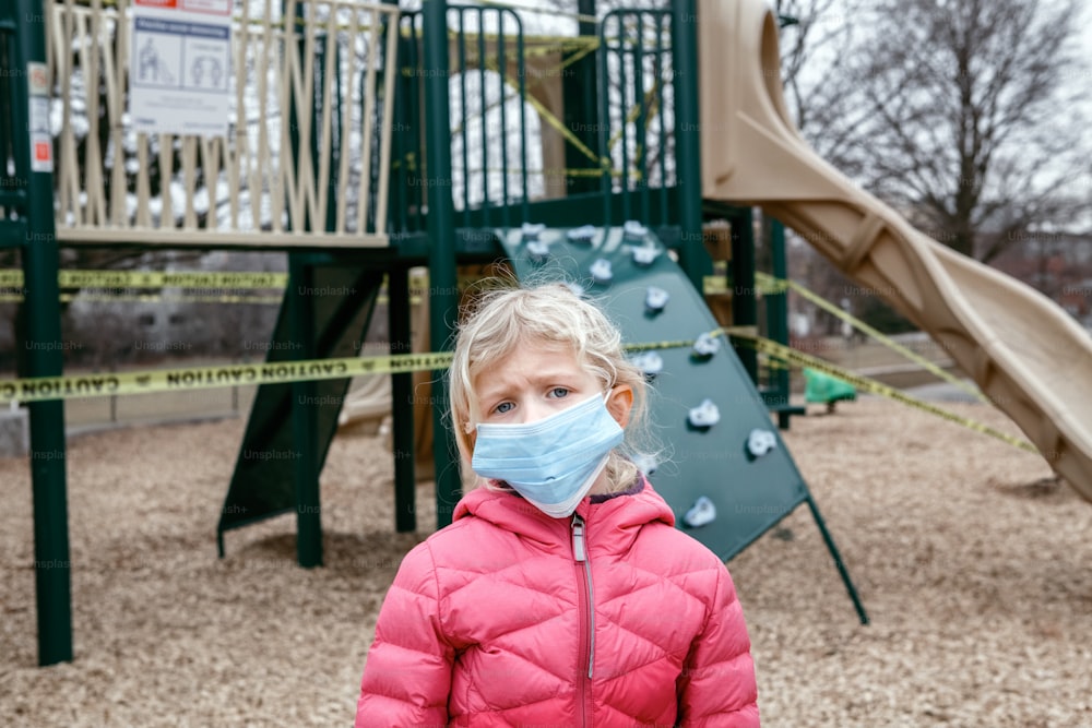 Portrait of sad Caucasian girl in face mask on closed playground outdoor. Kids play area locked with yellow caution tape in Toronto Canada. Coronavirus social distance quarantine.