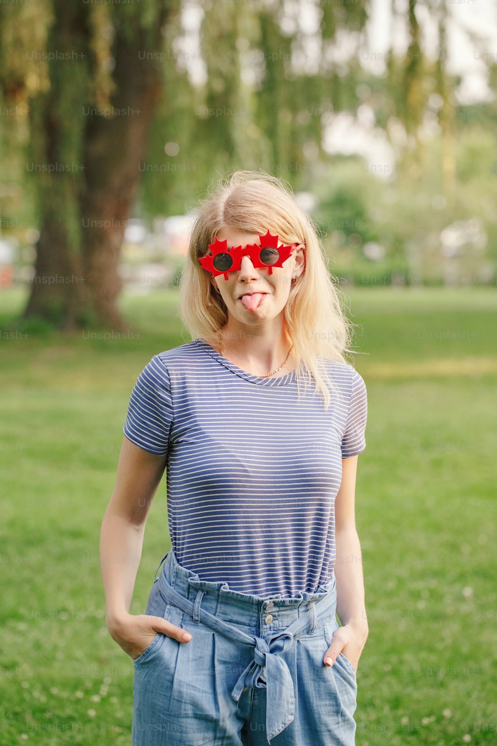 Funny blonde Caucasian young girl wearing canadian maple leaf sunglasses. Happy hipster woman in red sunglasses having fun outdoor. Citizen female celebrating Canada Day holiday on July 1.
