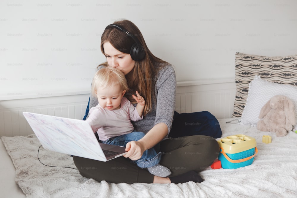 Young Caucasian mother with toddler baby working on laptop from home. Workplace of freelance woman student with kid. Online learning education or work job during self isolation, quarantine.